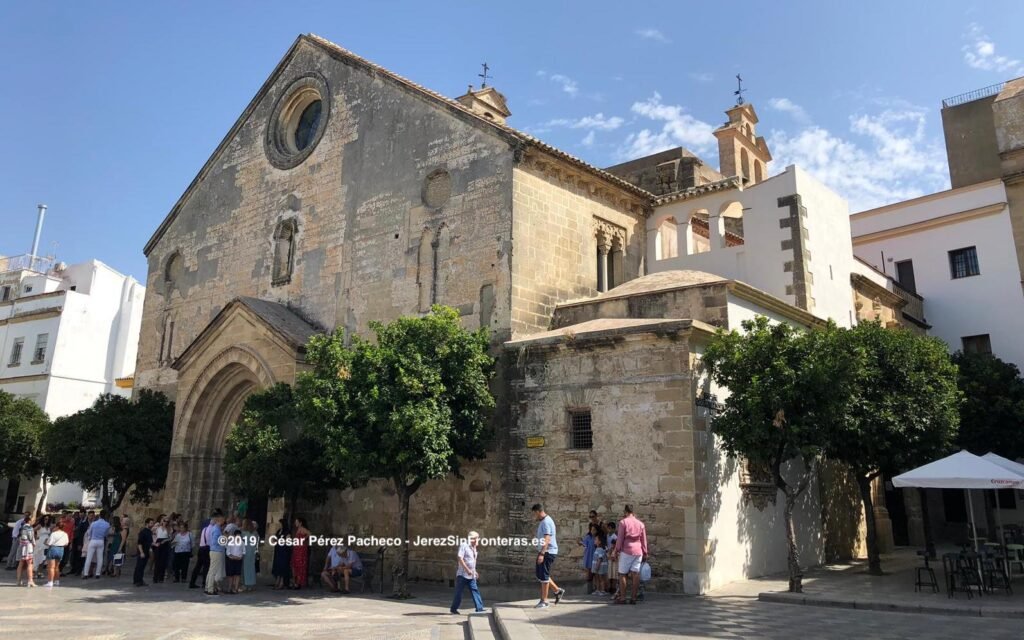 Fachada de piedra de la Iglesia de San Dionisio en Jerez, con personas esperando en la entrada y naranjos en la plaza.
