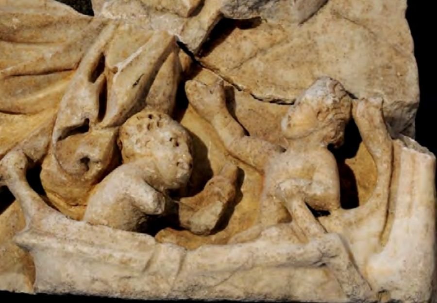 Detalle en primer plano de un fragmento de mármol del sarcófago de Medina Sidonia que representa a dos pequeños erotes esculpidos a bordo de una barca, remando sobre el oleaje marino tallado en relieve.