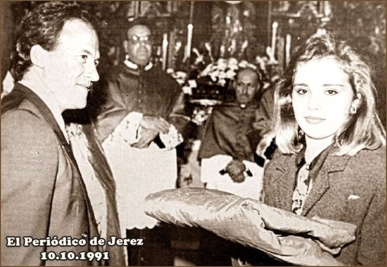 Fotografía histórica restaurada digitalmente y coloreada que muestra a cinco personas en el interior de una iglesia. A la izquierda, un hombre de perfil con traje oscuro y camisa blanca mira hacia la derecha. Detrás de él, dos clérigos con túnicas eclesiásticas, uno de ellos con birrete y gafas, y otro sentado, observan la escena. A la derecha, una mujer joven con una chaqueta de lana verde y cabello rubio corto mira hacia abajo con atención el gran fajo de tela de color rosa coral que sostiene en sus manos. La imagen está enmarcada por un borde marrón. En la esquina inferior izquierda, el texto "El Periódico de Jerez" arriba y "10.10.1991" abajo. El fajo de tela representa el Pendón de Jerez.