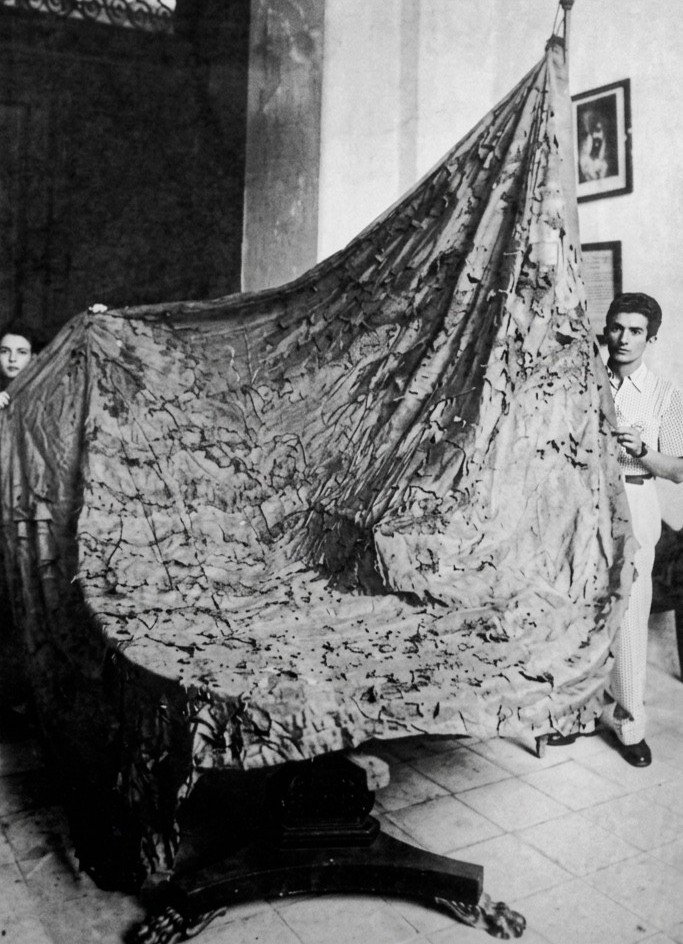 Fotografía histórica en blanco y negro, restaurada digitalmente, que muestra el antiguo Pendón de Jerez en un estado de conservación muy deteriorado y frágil. La gran tela medieval aparece desplegada verticalmente, llena de rasgones y zurcidos, sostenida en los extremos por dos personas dentro de una estancia de época con suelo de baldosas y un mueble de madera de estilo clásico en la parte inferior.