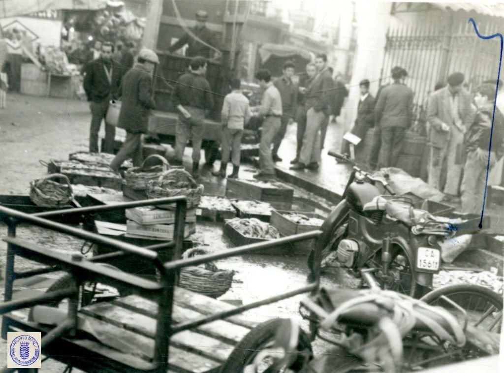 Fotografía histórica del abastecimiento de la Plaza de Abastos de Jerez en 1962, mostrando canastos de mimbre y cajas de madera con pescado en el suelo de la calle Doña Blanca, junto a una motocicleta y carretas de mano.