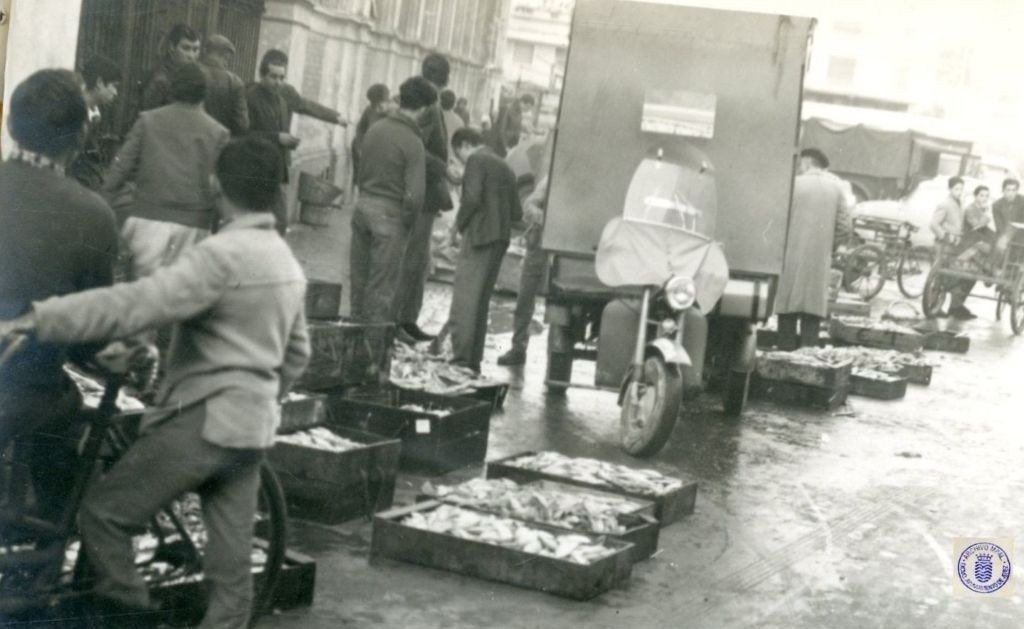 Fotografía en blanco y negro de la calle Doña Blanca en Jerez, 1962, capturando a un hombre en primer plano sobre una bicicleta, rodeado de cajas de pescado apiladas en el suelo mojado y un camión de reparto con el motor expuesto. Al fondo, un grupo de hombres observa la actividad frente a edificios del centro.
