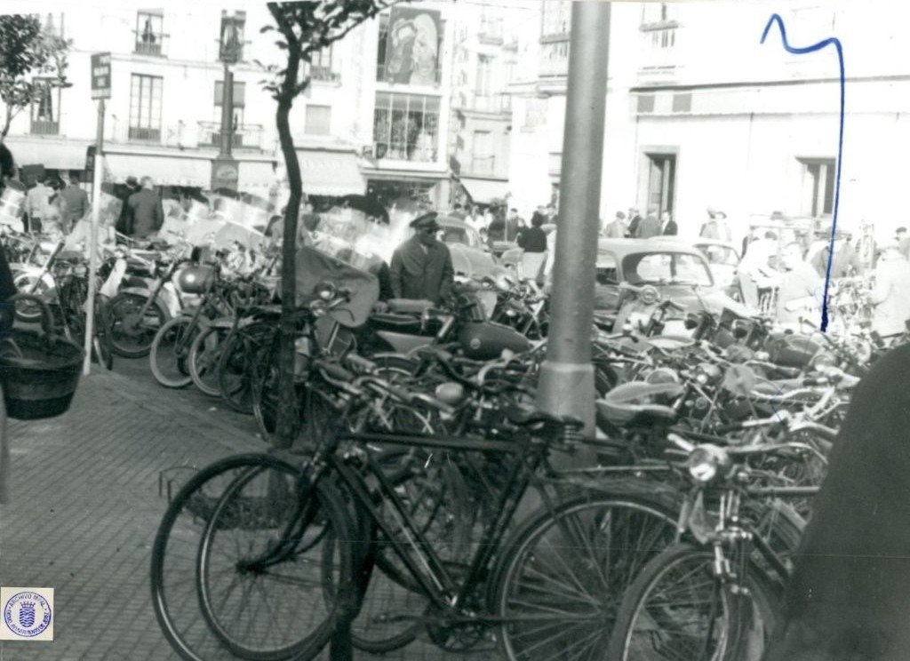 Fotografía en blanco y negro de 1962 que muestra una gran cantidad de bicicletas y motocicletas aparcadas en batería en la Plaza Esteve, junto a la calle Doña Blanca en Jerez. Al fondo se ven coches de la época y edificios de viviendas.
