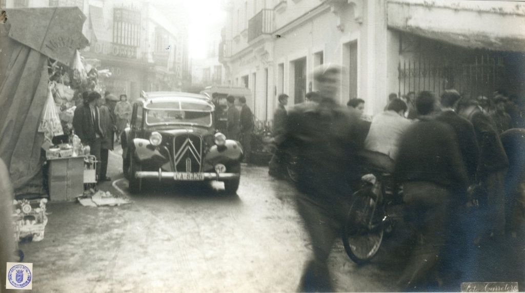 Un coche Citroën 11 Ligero (conocido como "Pato") con matrícula CA-8426 circulando por la calle Doña Blanca en 1962, rodeado de peatones y puestos de venta ambulante en un día de gran actividad comercial.