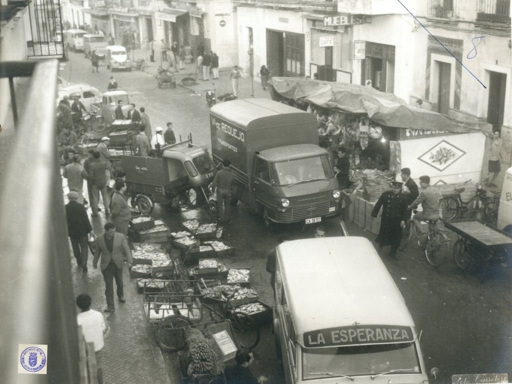 Vista elevada de la calle Doña Blanca en 1962 con furgonetas de "La Esperanza" y "Vda. de Requejo" maniobrando entre cajas de pescado por el suelo y puestos de venta ambulante.
