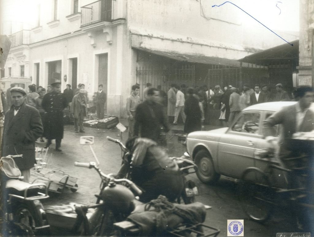 Fotografía en blanco y negro de 1962 que muestra la calle Doña Blanca en Jerez llena de gente. En primer plano, una motocicleta aparcada; en el centro, peatones y un coche en movimiento que aparece algo borroso, capturando el ajetreo urbano. Al fondo, edificios y el Mercado de Abastos.
