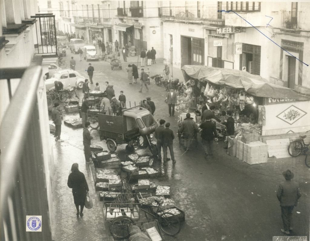 Fotografía histórica de la calle Doña Blanca en Jerez (1962), vista desde un balcón, mostrando un motocarro de reparto junto a decenas de cajas de pescado en el suelo y un puesto de turrones o dulces con toldo.