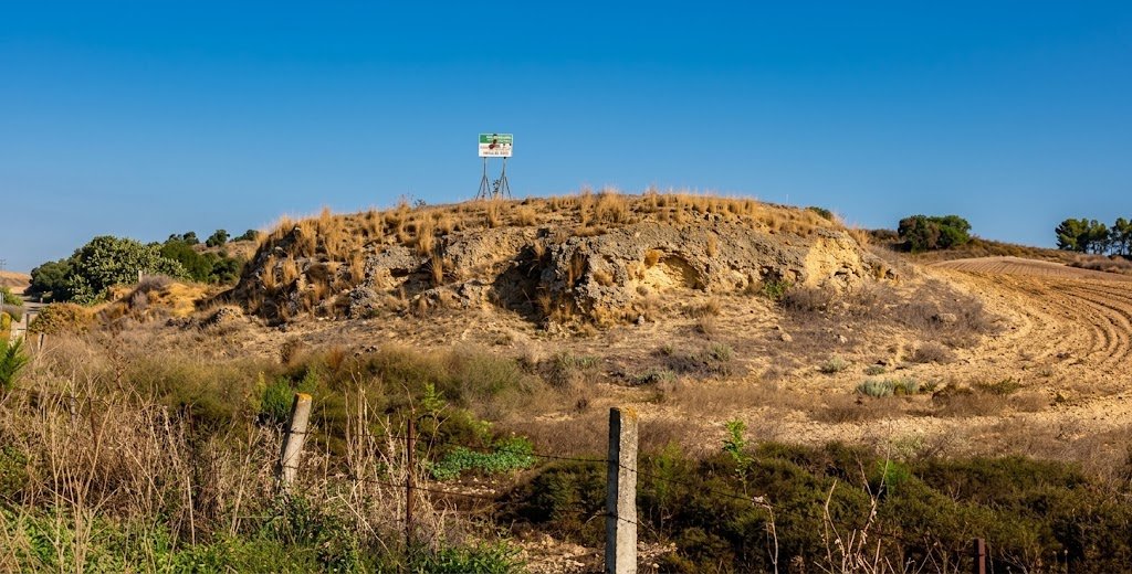 Panorámica del cerro y la elevación del yacimiento arqueológico de Asta Regia en la campiña de Jerez de la Frontera, con vegetación seca, terrenos agrícolas y la cartelería señalizadora en la cima bajo un cielo azul.