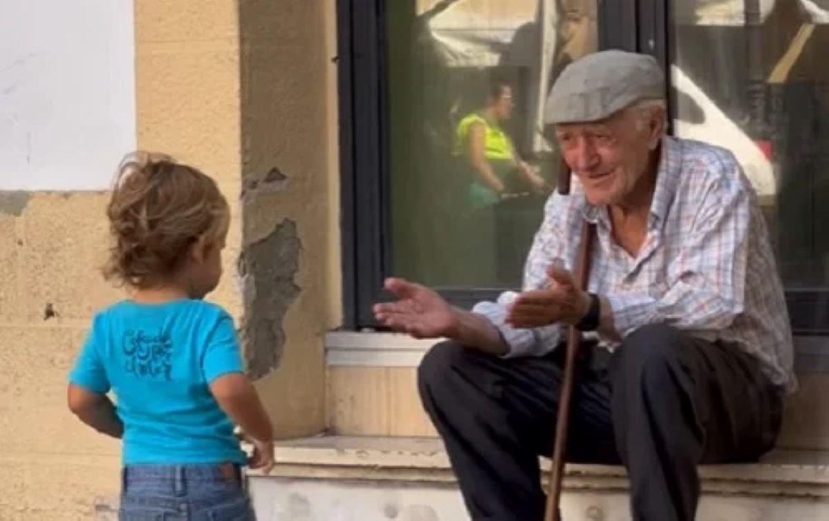 Anciano sentado en una calle de Jerez abre sus brazos sonriendo hacia un niño pequeño con camiseta azul.