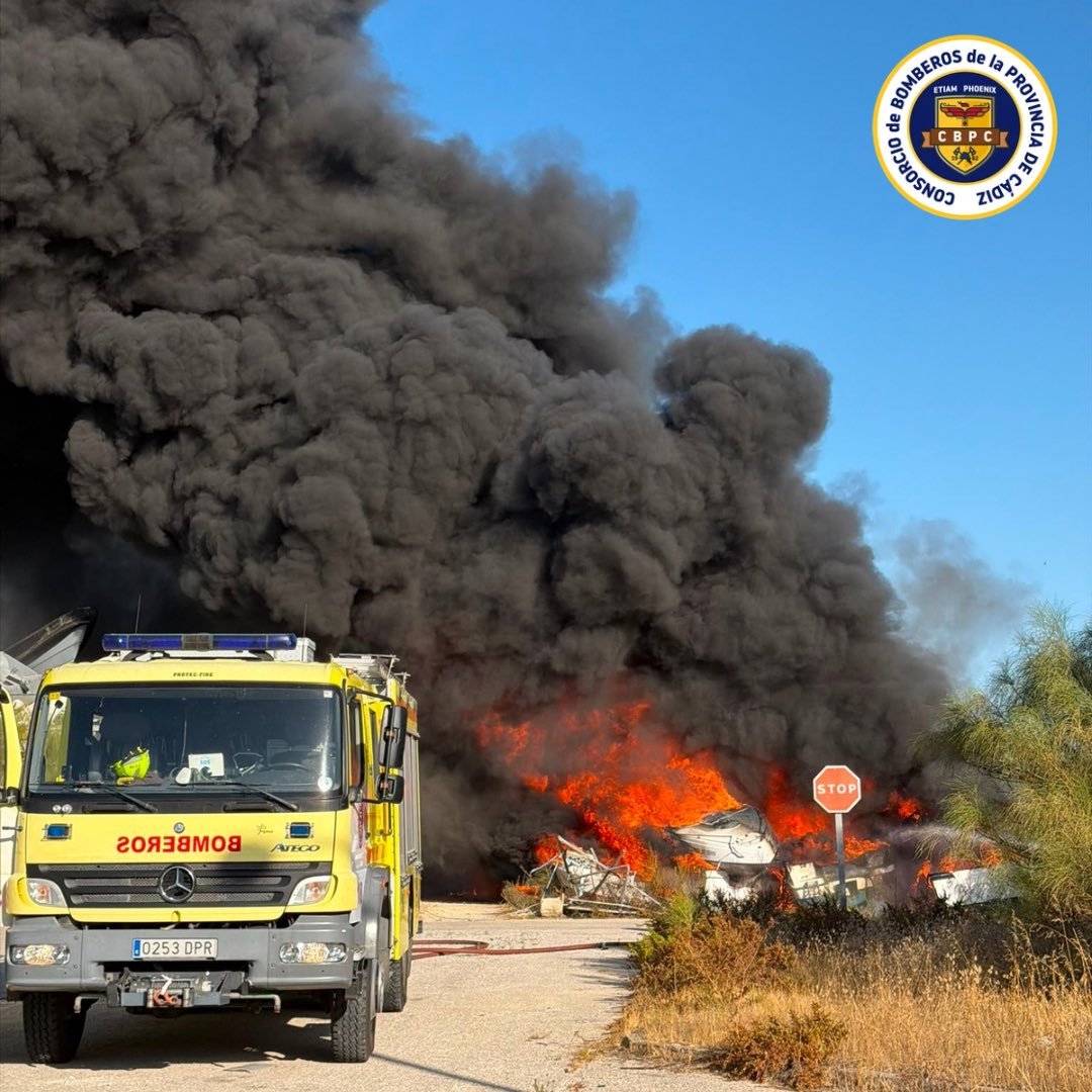 Camión de bomberos desplegado ante una densa columna de humo negro y llamas violentas en el depósito de embarcaciones del puerto.