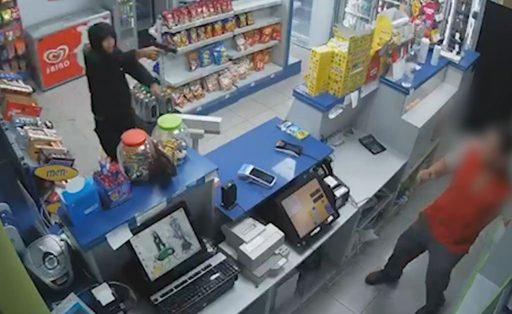Imagen de cámara de seguridad que muestra a un joven atracador encapuchado y vestido de negro, empuñando una pistola detrás del mostrador de una tienda de gasolinera. Un empleado con chaleco rojo está de pie frente al mostrador. El entorno de la tienda es visible, con estanterías y equipos de caja. El empleado está borroso para proteger su identidad.