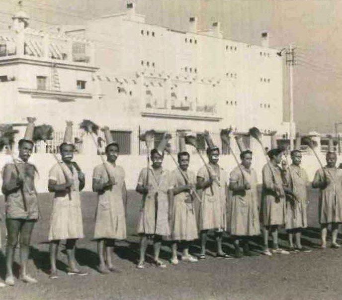 Fotografía antigua en blanco y negro de un grupo de hombres vestidos con trajes de sirvienta o enfermera de época y cofias, parados en línea en un patio. Cada uno sostiene una escoba de mango largo en el hombro como si fuera un rifle. Al fondo hay un gran edificio blanco de varios pisos con pancartas colgando.