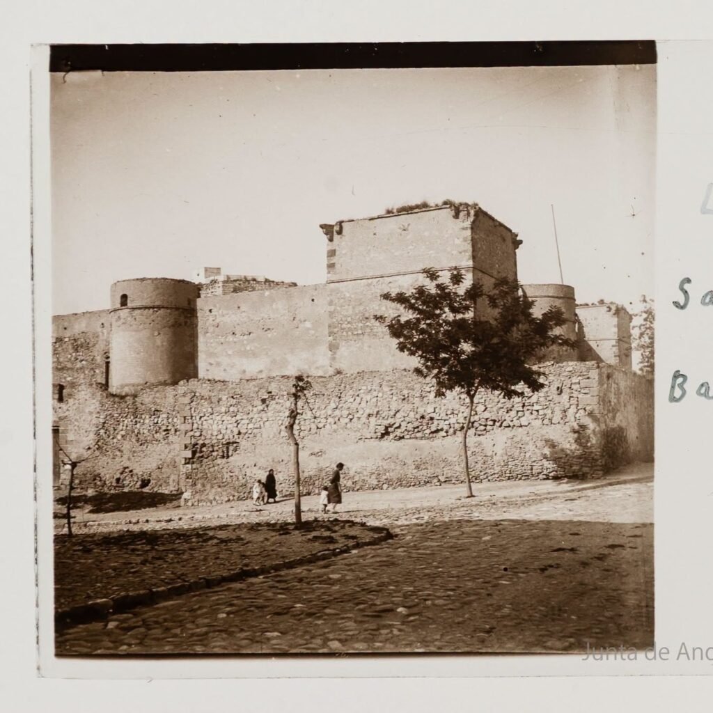 Fotografía en blanco y negro del Castillo de Santiago en Sanlúcar de Barrameda, con sus murallas de piedra y personas caminando por los alrededores.