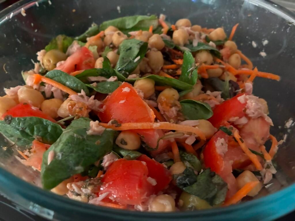 Ensalada fresca de garbanzos con atún desmenuzado, trozos de tomate natural, zanahoria rallada, aceitunas y brotes verdes en un bol transparente de cristal.