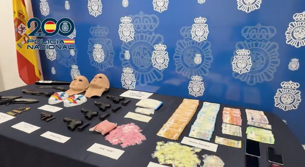 Fotografía de una mesa de exhibición de pruebas policiales con objetos incautados: máscaras faciales, armas cortas, dinero en efectivo (billetes de euro), bolsas con pastillas y polvo rosa, etiquetas identificativas y fondo con el escudo de la Policía Nacional y logo del 200 aniversario.