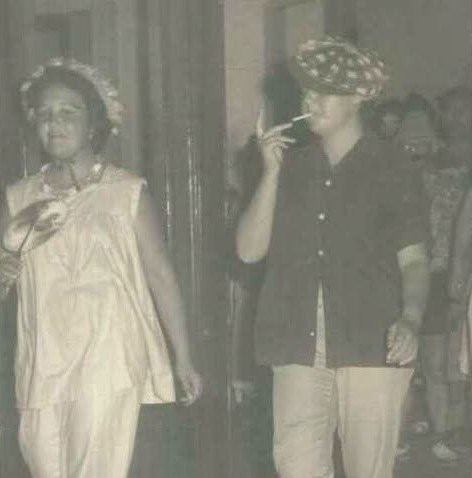 Fotografía en blanco y negro de los años 60 en la que aparecen dos personas caracterizadas en una de las jornadas festivas de la residencia gaditana. A la izquierda, una mujer sonríe con un vestido ligero y un tocado floral; a la derecha, otra persona con boina y camisa oscura fuma un cigarrillo mientras posa.
