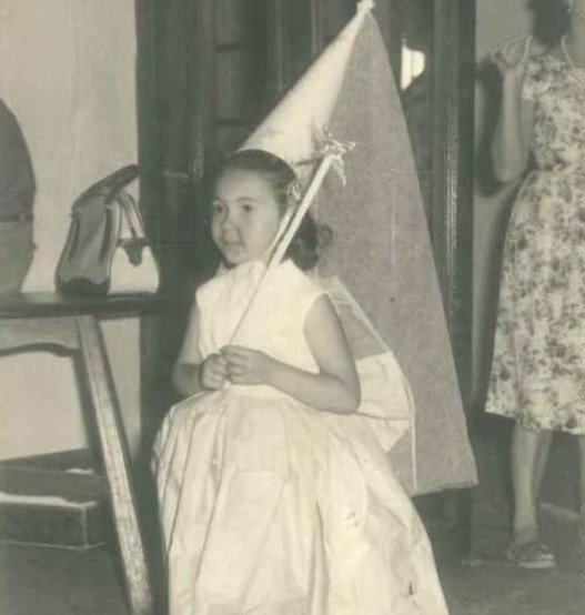 Fotografía en blanco y negro de los años 60 que muestra a una niña pequeña disfrazada con un vestido blanco y un sombrero cónico alto tipo capirote o gorro festivo, sosteniendo una pequeña varita decorada. De fondo se aprecia el interior de una estancia con muebles y una persona de pie con vestido estampado.