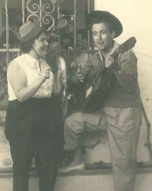 Fotografía vintage en blanco y negro que muestra a un hombre con sombrero cordobés tocando la guitarra junto a una mujer con sombrero de paja que toca la pandereta. Ambos sonríen y están vestidos con ropa tradicional o festiva frente a una reja de forja.