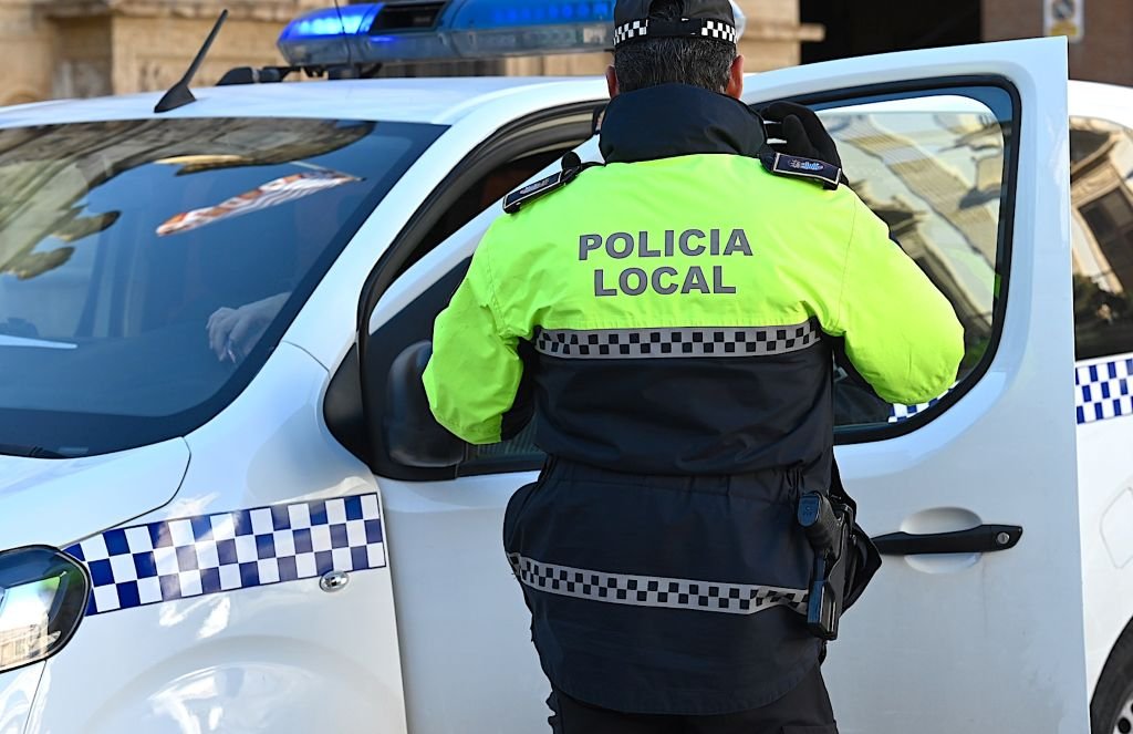 Un oficial de la Policía Local con una chaqueta amarilla de alta visibilidad de espaldas, parado junto a un coche de policía blanco con luces de emergencia azules, que tiene la puerta del pasajero abierta. El oficial tiene un arma de fuego visible en la cintura.