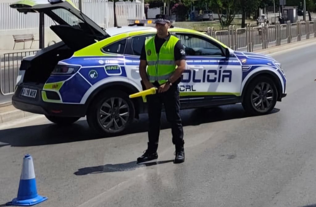 Un agente de la Policía Local de Jerez con chaleco reflectante realiza un control de tráfico en la calle junto a un coche patrulla con el maletero abierto.