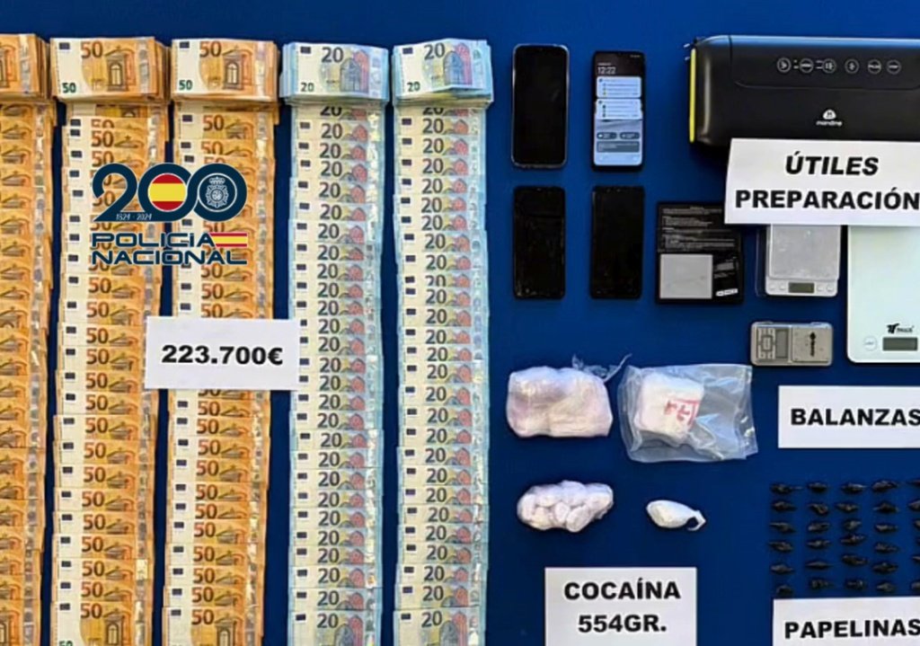 Fotografía oficial de la Policía Nacional mostrando una incautación de drogas y dinero en efectivo dispuestos sobre una mesa azul. Se ven fardos de billetes de 50 y 20 euros, 554 gramos de cocaína en polvo, papelinas envueltas individualmente, balanzas de precisión, un teléfono móvil y utensilios de preparación.