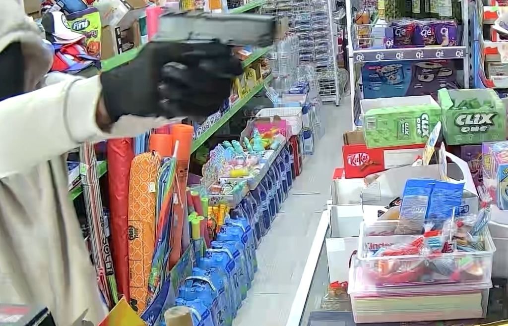 Primer plano de la mano con guante negro de un sospechoso sosteniendo una pistola plateada en la esquina inferior izquierda, apuntando hacia un pasillo de tienda de comestibles bien iluminado con estantes llenos de diversos productos y artículos para el hogar.