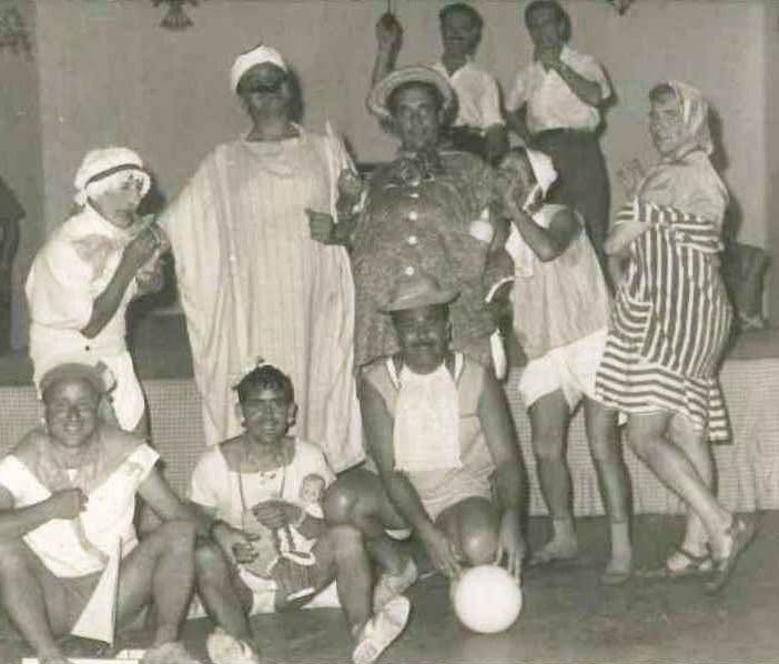 Fotografía en blanco y negro de los años 60 que muestra a un grupo de personas disfrazadas de forma cómica sobre un escenario durante una velada festiva. Llevan atuendos variados que incluyen túnicas, vestidos a rayas, sombreros y baberos, mientras posan sonrientes en distintas posturas, de pie y sentadas en el suelo.