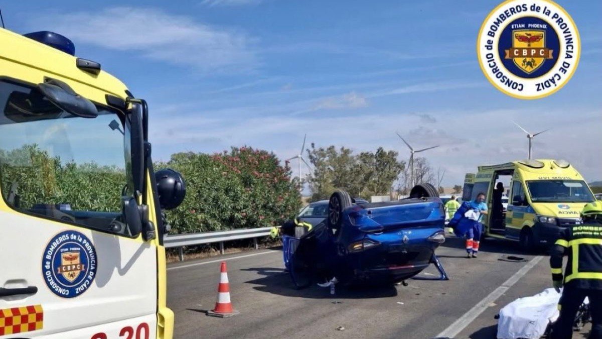 Fotografía que muestra la escena de un accidente de tráfico en una carretera. Un coche azul está volcado sobre su techo en el centro de la imagen. A la izquierda, el lateral de un camión de bomberos con la inscripción "S-20" y el logotipo del Consorcio de Bomberos de la Provincia de Cádiz. Al fondo, a la derecha, una ambulancia y varios bomberos atendiendo a una persona en una camilla.
