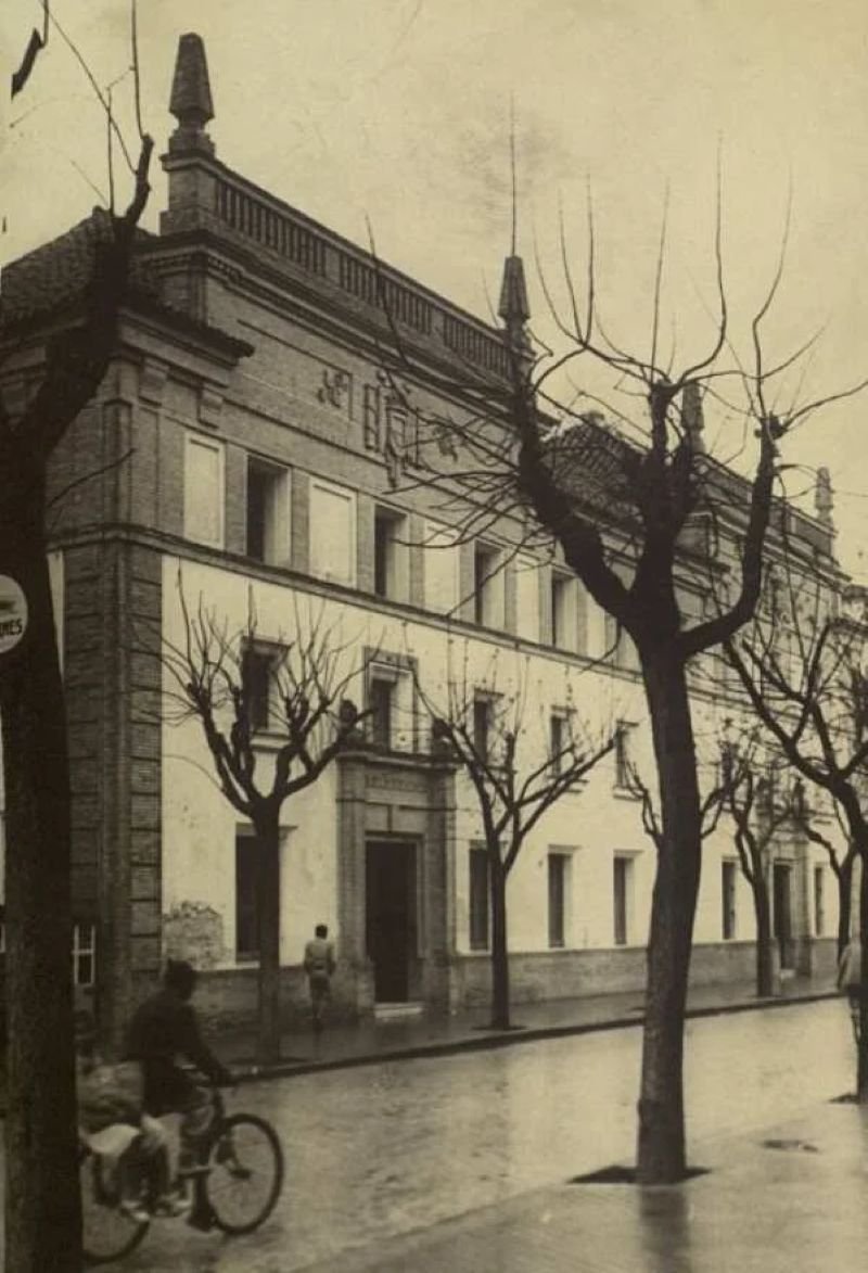 Fotografía histórica en blanco y negro que muestra la fachada principal de la Escuela de Arte y Superior de Diseño de Jerez. El edificio, de estilo regionalista, destaca por su combinación de ladrillo visto en la planta superior y paramentos enlucidos en la inferior, con una portada principal de piedra labrada. La imagen captura la calle con pavimento mojado y una hilera de árboles plátanos sin hojas. En primer plano, se distinguen figuras humanas: un ciclista en movimiento y un peatón caminando frente a la entrada.
