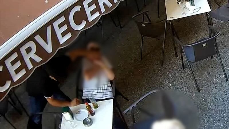 Una toma de seguridad aérea muestra una terraza de café vacía. Las mesas y sillas están ordenadas, con ceniceros en la mayoría. En primer plano, una mesa tiene algunas botellas y un pequeño cenicero de metal. En el borde izquierdo, el final de un letrero de neón que dice "...CERVECE" es visible. La escena está en un ángulo que muestra las filas de asientos que retroceden hacia la distancia.