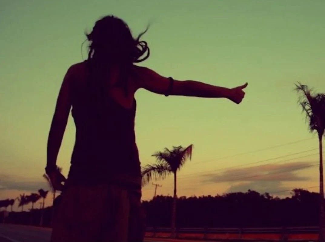 Silueta de Lorena Kej en una carretera al atardecer, con el brazo extendido haciendo autostop, simbolizando el viaje espiritual de su versión de Semilla Negra y el movimiento Nomadanz.