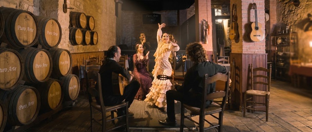 Una bailaora flamenca en el centro del escenario con un vestido de lunares voluminosos de color crema, con los brazos en alto. Un guitarrista está sentado a la izquierda y otro a la derecha, de espaldas. Una cantaora sentada en el fondo canta y aplaude. Detrás, otros artistas están de pie. El escenario está en una sala tradicional con paredes de piedra y vigas de madera. En el fondo, hay una pared de ladrillo visto y un estante con botellas. Una señal de "Puro Arte" es visible.