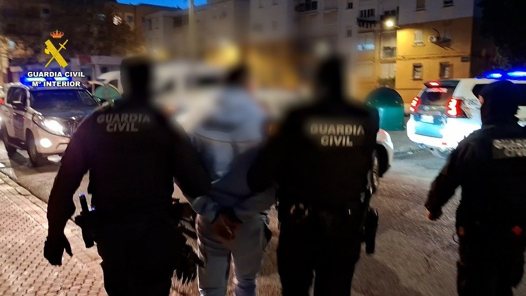 Dos agentes de la Guardia Civil escoltan a un detenido esposado durante la noche en una calle de la provincia de Cádiz, con vehículos policiales de fondo.