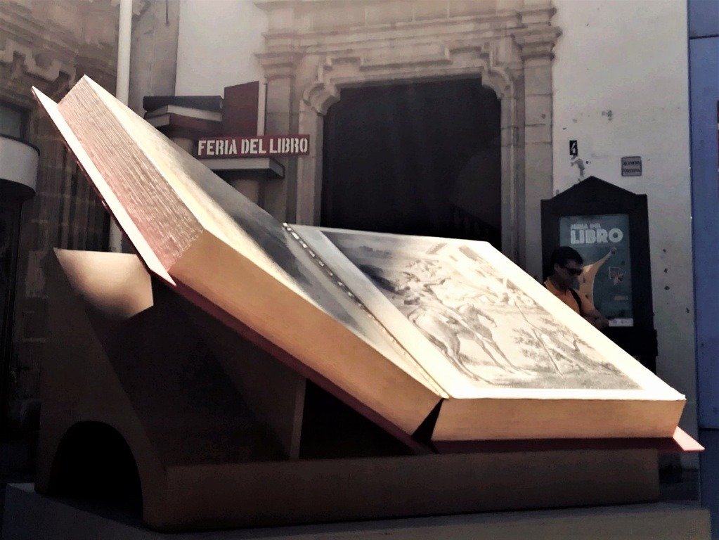 Escultura monumental de un libro abierto con ilustraciones clásicas frente a la entrada de la Feria del Libro de Jerez, destacando la temática de cultura y literatura.