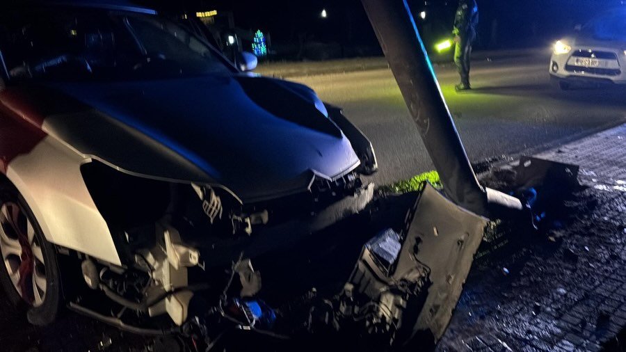 Plano detalle del frontal destrozado de un coche accidentado en Medina Sidonia, con un agente de policía regulando el tráfico con una linterna al fondo.
