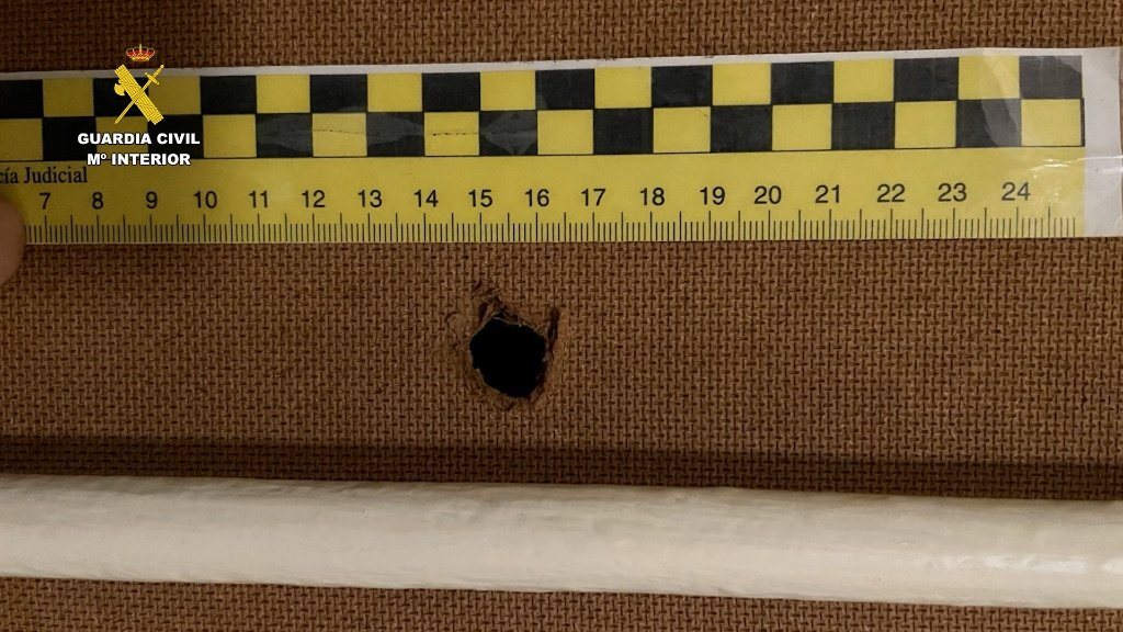 Primer plano de un orificio circular realizado en un panel de madera, medido con una regla pericial de la Guardia Civil.