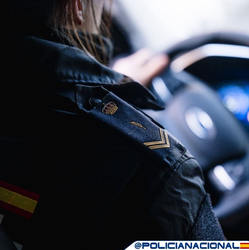 Primer plano de un agente de la Policía Nacional al volante de un coche patrulla, mostrando el parche de la bandera de España en el uniforme.