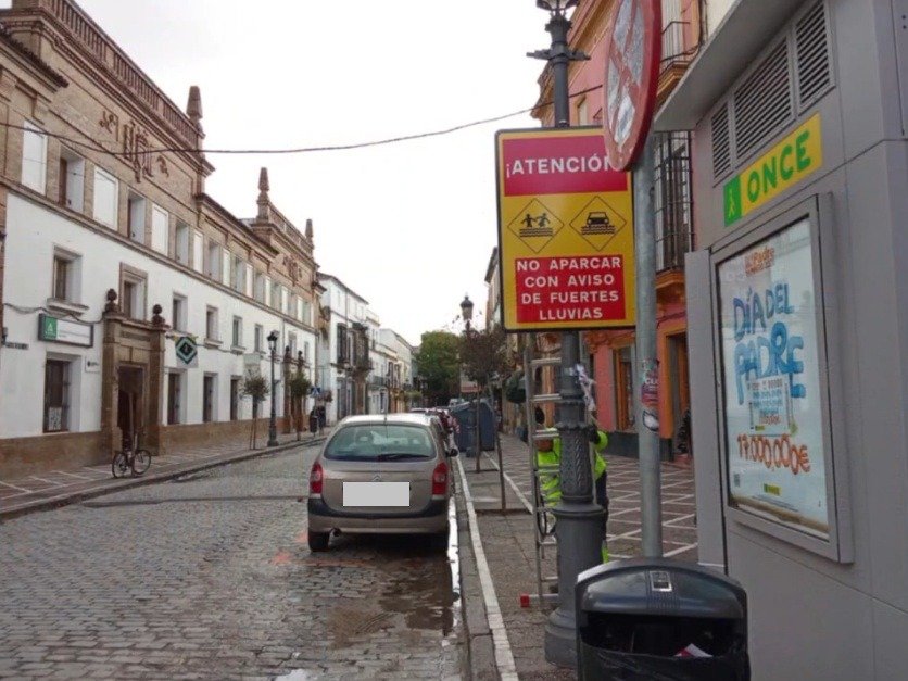Plano medio de señal roja y amarilla con aviso de no aparcar por lluvias intensas en una calle con pavimento de adoquines y arquitectura tradicional en Jerez.
