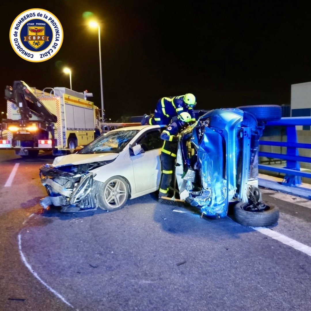 Accidente nocturno en Puerto Real con un coche blanco destrozado y uno azul volcado; bomberos trabajando con camión de emergencias al fondo.