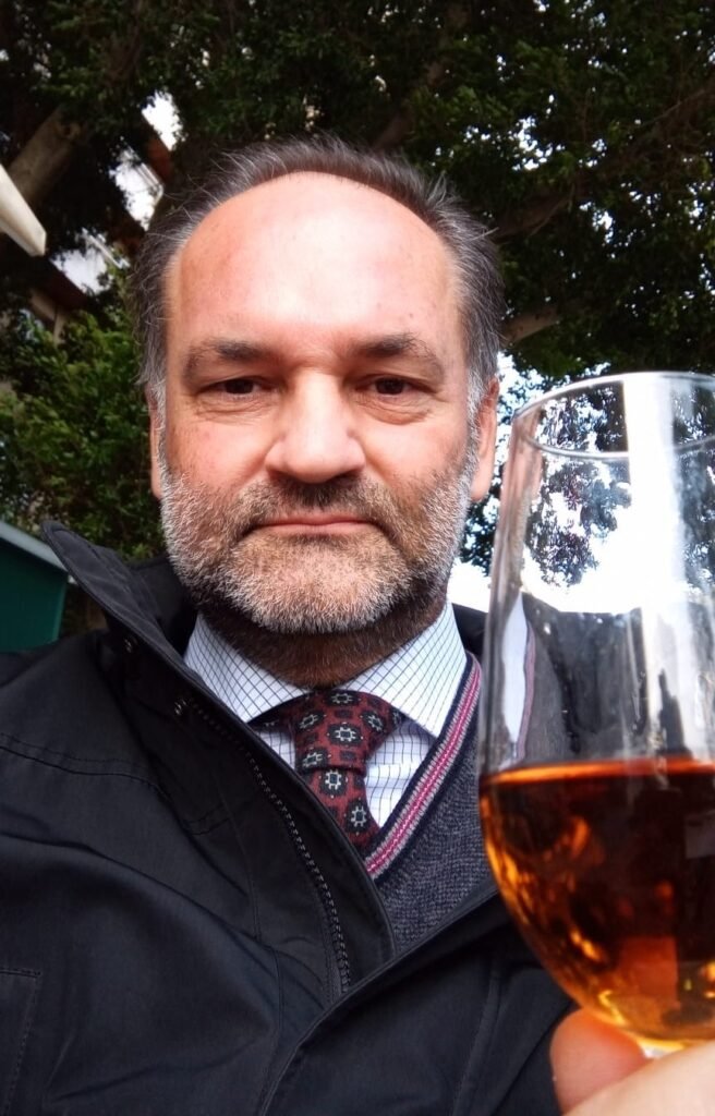 Retrato en primer plano de Bosco Gallardo Quirós en Jerez de la Frontera, sonriendo levemente y sosteniendo una copa de vino generoso ambarino frente a unos árboles de fondo.