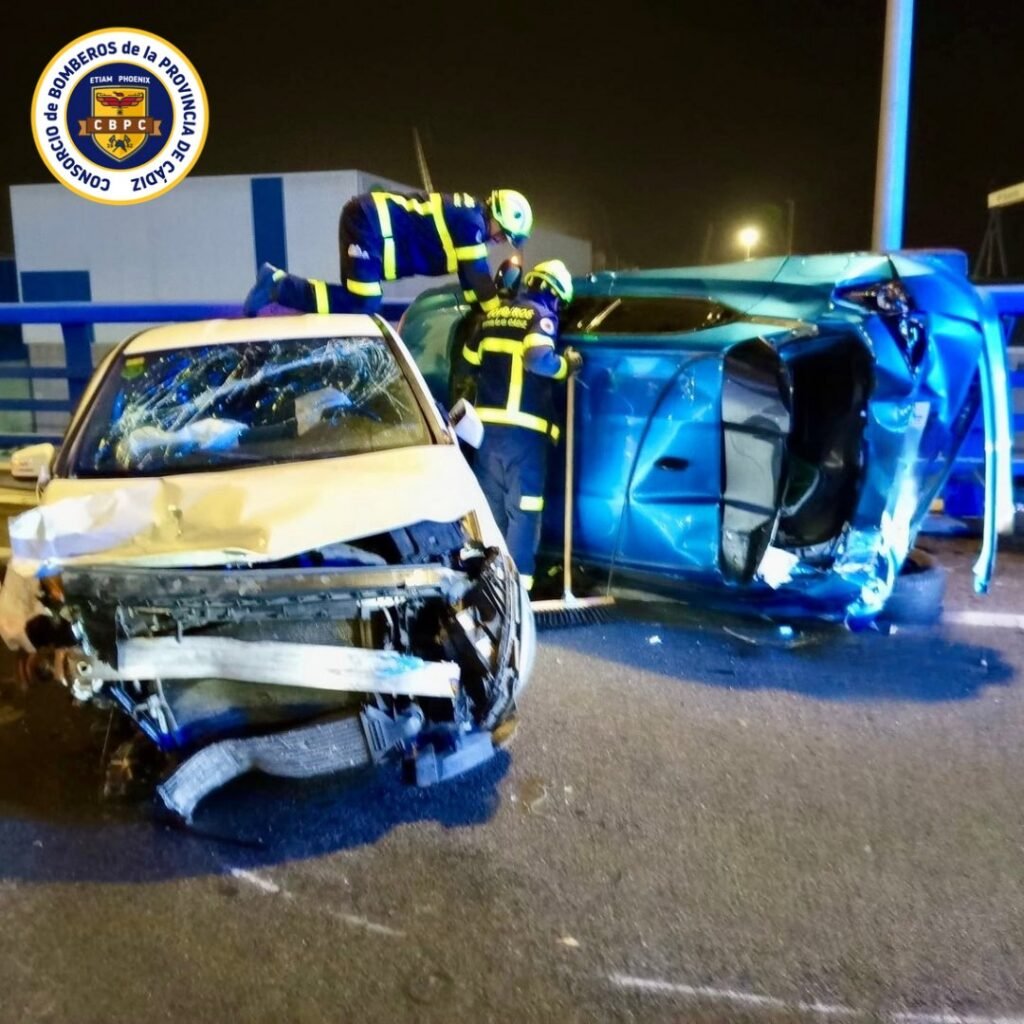 Frontal de turismo blanco completamente aplastado tras choque frontal en Puerto Real; operarios limpian la vía al fondo.