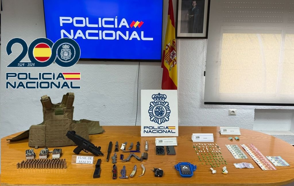 Fotografía de una mesa oficial de la Policía Nacional mostrando todo el material incautado: un fusil de asalto, munición, navajas, un chaleco antibalas, numerosas dosis de cocaína en bolsitas verdes, fajos de billetes de euro y teléfonos móviles. Al fondo, un televisor con el logo de la Policía Nacional y la bandera de España.