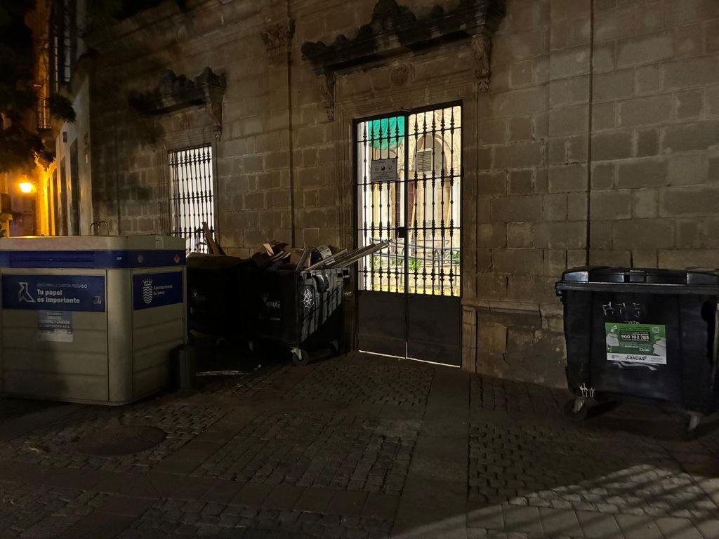 Zona de contenedores nocturnos en la Plaza del Progreso junto a un portal con rejas.
