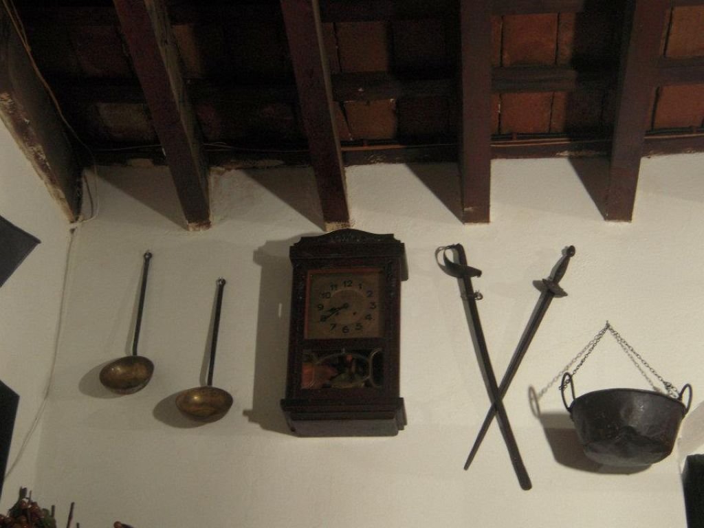 Reloj de pared antiguo flanqueado por dos espadas cruzadas y calderos de cobre en una pared blanca.