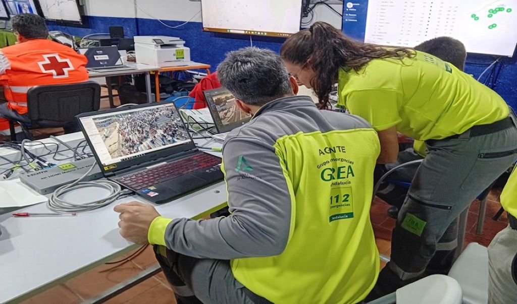 Primer plano de espaldas de un técnico del Grupo de Emergencias de Andalucía (GREA) con chaqueta amarilla reflectante, consultando una pantalla de ordenador portátil. A su lado, otra técnico con polo amarillo reflectante y gafas de sol en la cabeza mira la pantalla. En el fondo, se ve a otra persona con chaleco de Cruz Roja y varias pantallas grandes con mapas y datos en una pared azul. El entorno es una sala de control de emergencias tecnológica.