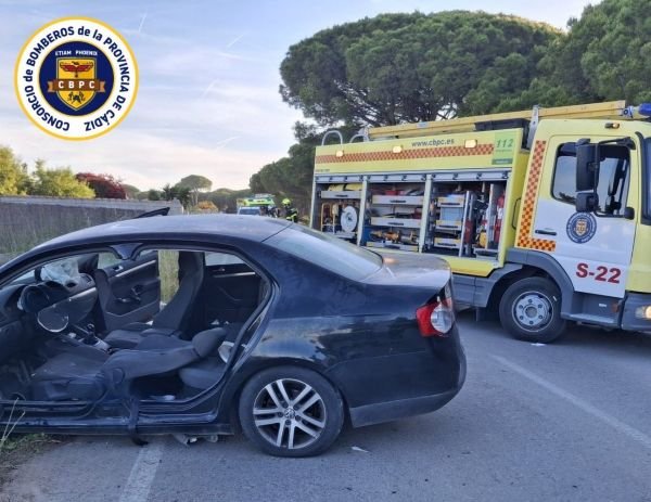 Coche de color negro con graves daños estructurales en el lateral izquierdo tras sufrir una salida de vía en Conil de la Frontera, junto a un camión de rescate del Consorcio de Bomberos de la Provincia de Cádiz.