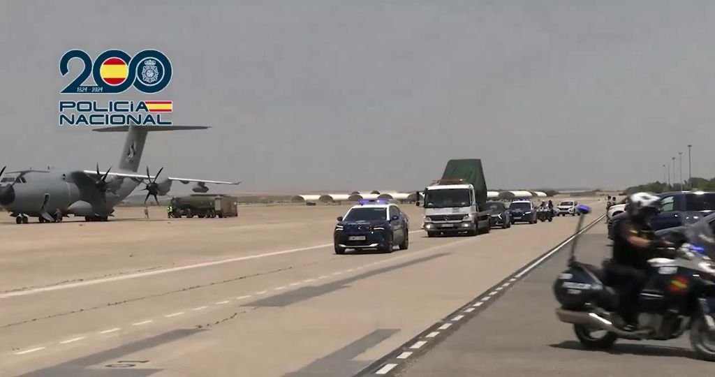 Convoy de la Policía Nacional escoltando un camión con el papamóvil cubierto por una lona en una pista militar al lado de un avión del Ejército del Aire.