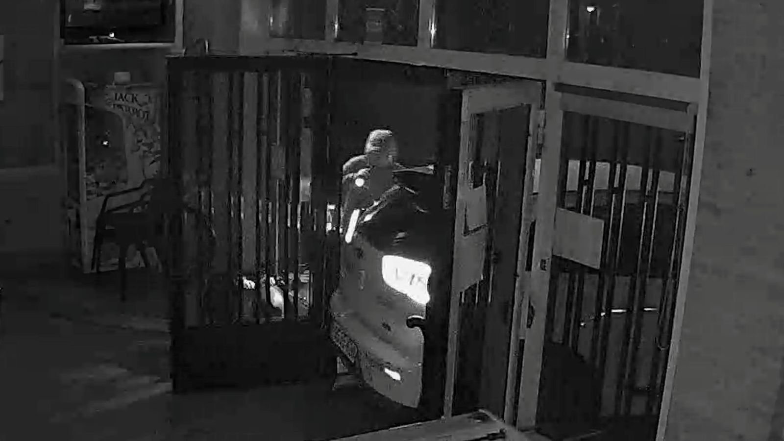 Captura de cámara de seguridad nocturna en blanco y negro que muestra un coche blanco empotrado en la entrada destrozada de un establecimiento comercial durante un robo por alunizaje.
