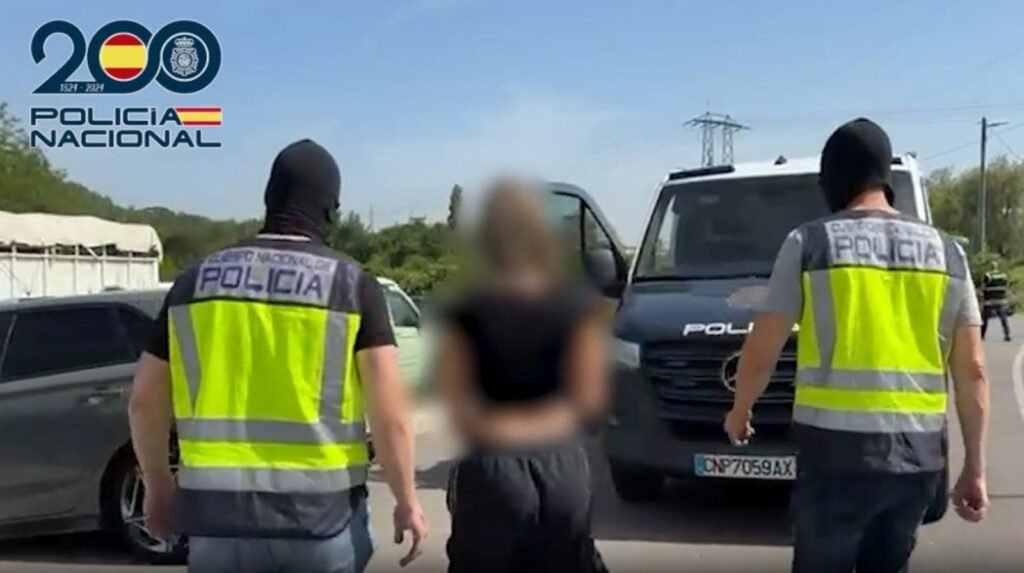 Dos agentes de la Policía Nacional escoltan de espaldas a un detenido esposado hacia un furgón policial durante el día.