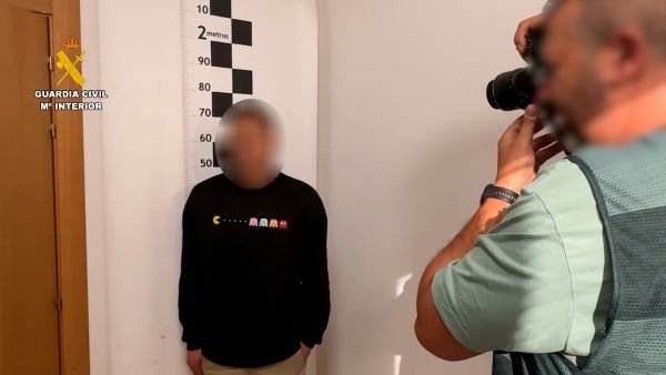 Un agente de espaldas fotografía a un hombre detenido con el rostro pixelado frente a un medidor de estatura en las dependencias de la Guardia Civil.